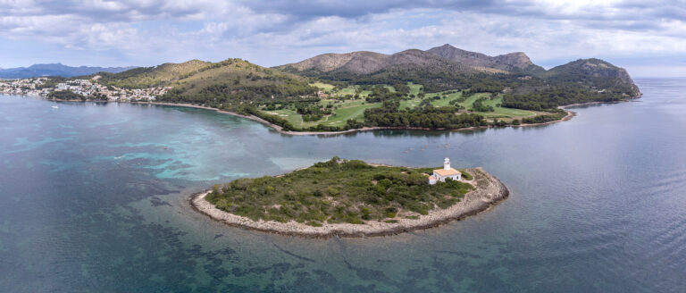 Vista aérea del islote de Alcanada y la bahía de Alcudia Fotografía aérea panorámica del islote de Alcanada con su faro, el campo de golf y la costa de Alcudia al fondo sobre aguas turquesas.