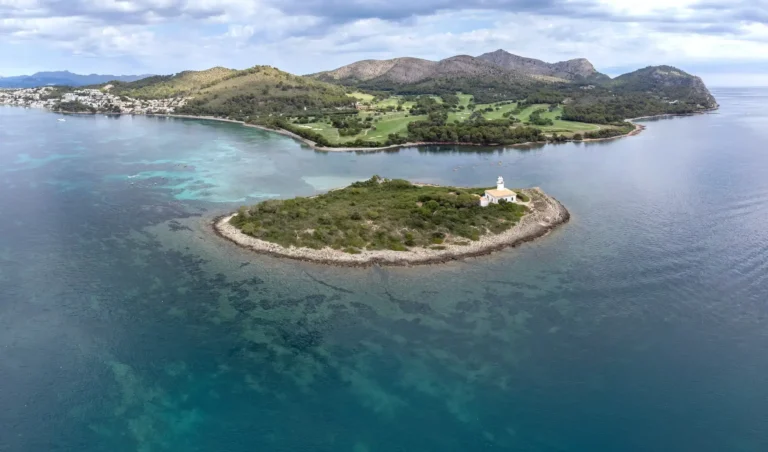 Islote de Alcanada con el Faro de Alcanada, Bahía de Alcúdia, Mallorca Vista aérea del Islote de Alcanada con el Faro de Alcanada en su centro rodeado de aguas transparentes de la Bahía de Alcúdia y pinar en el islote, Mallorca